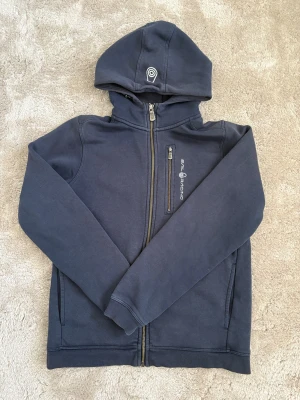 Mörkblå hoodie från Sail Racing - Mörkblå hoodie från Sail Racing med dragkedja hela vägen, huva och en bröstficka med dragkedja. Snygg logga på bröstet och huvan. Mjuk bomullskänsla och ribbade muddar vid ärmslut och nederkant. Perfekt för chill dagar.