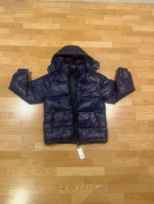 Mörkblå pufferjacka från Polo Ralph Lauren - Snygg mörkblå pufferjacka från Polo Ralph Lauren med röd broderad logga på bröstet. Jackan har huva, två sidofickor med dragkedja och RL-detaljer på zip. Materialet är glansigt syntet och jackan är quiltad för extra värme. Perfekt för kalla dagar.