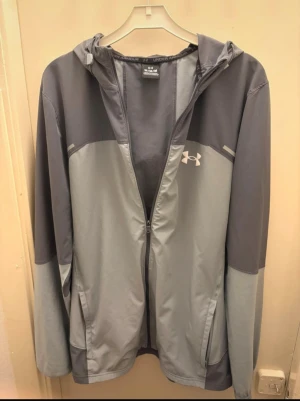 Grå vindjacka från Under Armour - Snygg tvåtonad grå vindjacka från Under Armour med huva och dragkedja framtill. Jackan har logga på bröstet och ett stort tryck på ryggen. Långärmad och tillverkad i lätt polyester, perfekt för aktiva dagar utomhus. Lite skada på armen men de syns inte efter som jag har sytt ihop den 