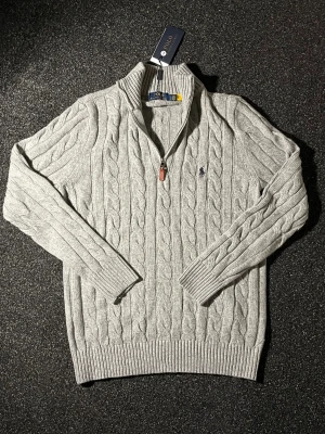 Grå stickad half zip Polo Ralph Lauren - Snygg grå stickad tröja från Polo Ralph Lauren i storlek L. Tröjan har ett klassiskt kabelstickat mönster, half zip med läderdetalj och ribbade muddar. Perfekt för dig som gillar stilren och tidlös look. Polo-logga broderad på bröstet.