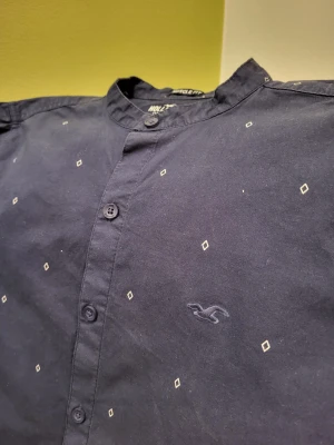 Muscle fit Mörkblå Hollister henley kortärmad - Kortärmad mörkblå henley-skjorta från Hollister med diskret diamantmönster och broderad logga på bröstet. Skjortan har muscle fit-passform, ståkrage och knappar framtill. Tillverkad i mjuk bomull med lite stretch för extra komfort.