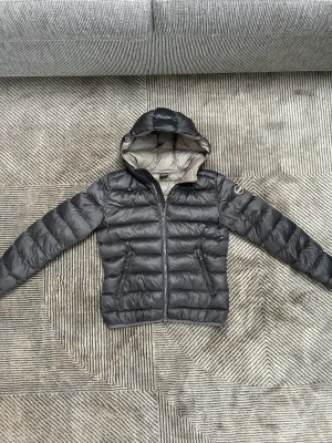 Mörkgrå dunjacka från Colmar - Snygg mörkgrå dunjacka från Colmar med huva och grått foder. Jackan har quiltad design, två sidofickor med dragkedja och Colmar-logga på ärmen. Perfekt för kalla dagar och har en modern, sportig look som funkar till streetwear.