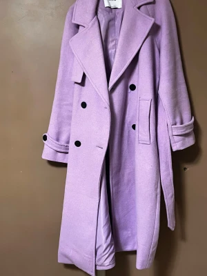 Ljuslila kappa från Bershka XS - Säljer en ljuslila kappa från Bershka i storlek XS. Kappan har dubbelknäppning med svarta knappar, bred krage, fickor och bälte i midjan. Materialet är mjukt och känns ulligt, med ett slätt innerfoder. Perfekt för dig som vill sticka ut i höst och vinter.