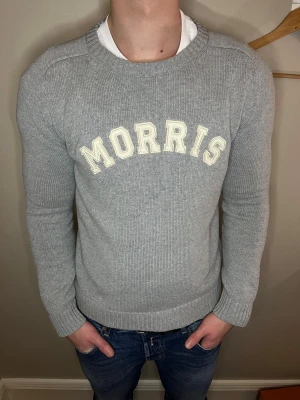 Grå stickad tröja från Morris - Snygg grå stickad tröja från Morris i storlek medium. Tröjan har ett klassiskt rund hals och vit text med Morris broderat på bröstet. Ribbstickade muddar vid ärmslut och nederkant ger en schysst passform. Perfekt för dig som gillar stilrena och bekväma plagg. Pris är diskuterbart vid snabb affär.