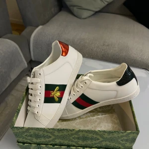 Gucci Ace Bee Sneakers i vitt skinn - Gucci Ace sneakers i vitt skinn med klassiska gröna och röda sidoband och broderad guldbi. Baksidan har metallicdetaljer i rött och grönt med ormskinnsstruktur och Gucci-logga. Skorna har vita snören och platt sula. Riktigt ikoniska och snygga sneakers för dig som gillar lyxig streetstyle.