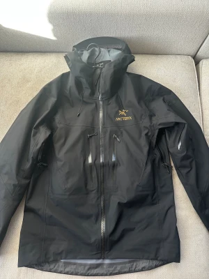 Arc'teryx jacka  - Arc'teryx svart vindjacka med gul broderad logga på bröstet, flera vattentäta dragkedjor och justerbar huva. Jackan är tillverkad i slitstark polyester och har RECCO-reflektor för extra säkerhet. Perfekt för dig som gillar outdoor och vill ha en snygg techjacka. Passar perfekt som skidjacka. Passar även storlek M. Passar både kille och tjej