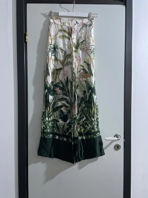 Mönstrade wide byxor från Vila - Snygga wide byxor från Vila med tropiskt mönster i grönt, vitt och inslag av rosa och beige. Byxorna har hög midja och croppad längd, tillverkade i ett mjukt och lätt material som känns skönt mot huden. Perfekta för dig som gillar att sticka ut med din stil.