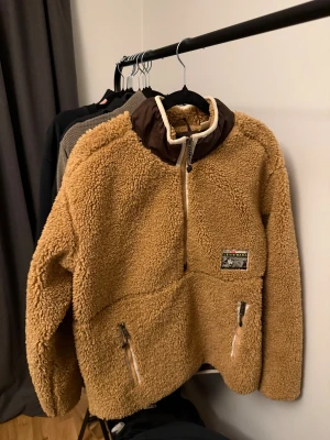 Levi's teddyjacka i beige S - Säljer en beige teddyjacka från Levi's med bruna detaljer och halv dragkedja. Jackan har mysigt fluffigt material, två fickor med dragkedja och coolt Levi's Warm-märke på bröstet. Perfekt för kalla dagar och chill stil.