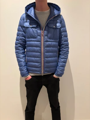 Blå Moncler dunjacka med huva - Säljer en blå dunjacka från Moncler med huva och quiltad design. Jackan har två bröstfickor med lock och knappar, samt dragkedja framtill, dragkedjan har några skavanker som vi kan fixa vid köp. Och med Monclers klassiska ränder. Perfekt för dig som vill ha en snygg och varm jacka med street vibe.