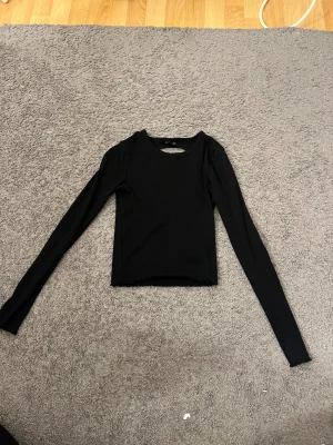 Svart ribbad långärmad topp med öppen rygg - Säljer en svart ribbad långärmad topp med en snygg öppen rygg. Toppen är tight i modellen och har en rundad halsringning framtill. Perfekt för dig som gillar stilrena basplagg med en twist.