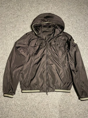 Svart vindjacka från Moncler - Svart vindjacka från Moncler med huva och dragkedja framtill. Jackan har två bröstfickor med knapp, ribbade muddar med ränder och Moncler-logga på ärmen. Materialet är glansigt men inte likadant som på mayan och jackan har snörning vid huvan för extra stil. Öppen till frågor, extra bilder och pris kan diskuteras.