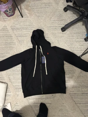 Svart hoodie från Polo Ralph Lauren - Svart hoodie med dragkedja från Polo Ralph Lauren. Har vit dragsko i huvan, klassisk röd Polo-logga broderad på bröstet och två fickor framtill. Mjuk bomullskänsla och stilren design som passar perfekt till jeans eller joggers.