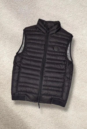 Svart pufferväst från Armani Exchange XS (ny) - Oanvänd  svart väst från Armani Exchange i storlek XS. Skulle säga att den även passar S! Västen har quiltad design, hög krage och dragkedja framtill. Insidan är ljusgrå och nederkanten har ett brett band med logotypen. Perfekt att slänga över en hoodie för extra värme.