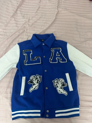 Blå och vit bomberjacka med LA och änglar - Snygg blå bomberjacka med vita ärmar och stora LA-bokstäver framtill. Jackan har broderade änglar både fram och bak, samt vita ränder på muddar och fickor. Stängs med tryckknappar och har klassisk college-stil. Perfekt för dig som vill sticka ut.