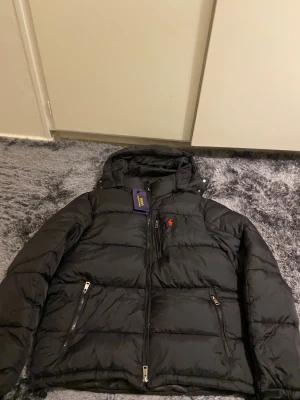 Svart pufferjacka från Polo Ralph Lauren - Svart pufferjacka från Polo Ralph Lauren med röd broderad logga på bröstet. Jackan har huva, dragkedja framtill och två sidofickor med dragkedja. Materialet är syntet och jackan har en quiltad design som ger en schysst look.