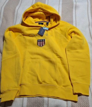 Gul hoodie från GANT med logga - Oanvänd Gul hoodie från GANT med broderad logga och amerikanskt inspirerat märke på bröstet. Klassisk känguruficka framtill och huva med dragsko. Mjuk bomullsblandning och ribbade muddar vid ärmslut och nederkant. Perfekt för en avslappnad och sportig stil.