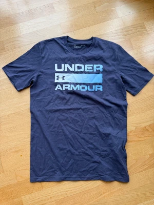 Blå Under Armour t-shirt i bomull - Snygg mörkblå t-shirt från Under Armour med stort tryck på bröstet. Modellen har rund hals och korta ärmar. Perfekt för dig som gillar sportiga och bekväma plagg med stilren design.