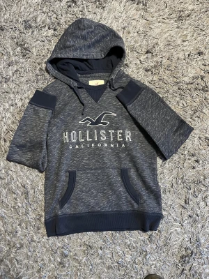 Mörkblå Hollister hoodie med kort ärm - Snygg mörkblå hoodie från Hollister med korta ärmar och vit logga framtill. Tröjan har huva med snörning, känguruficka och är tillverkad i mjuk bomullsblandning. Perfekt för chill dagar och streetwear-vibe.