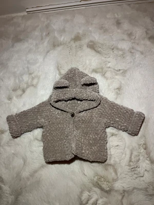 Beige teddyjacka med öron på luvan - Supermysig teddyjacka i beige med fluffig textur och huva med söta öron. Jackan har en knapp framtill och långa ärmar. Perfekt för att hålla sig varm och se gullig ut under kyliga dagar. För baby, nyfödda storlek 1-3 månader. 