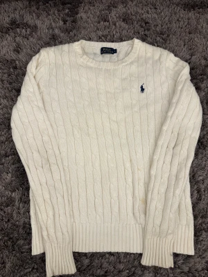 Vit kabelstickad tröja Polo Ralph Lauren - Snygg vit kabelstickad tröja från Polo Ralph Lauren i storlek S. Klassisk design med rund hals och ribbade muddar. Tröjan har det ikoniska blå broderade Polo-logot på bröstet och är långärmad. Perfekt för dig som gillar stilrena och tidlösa plagg.