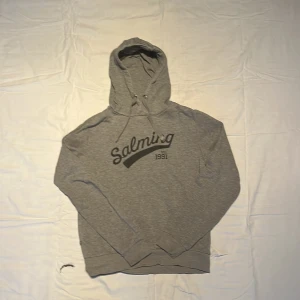 Grå hoodie från Salming med tryck - Grå hoodie från Salming med svart tryck på bröstet och snörning i huvan. Mjuk insida i fleece och klassisk passform. Perfekt för chill dagar eller när du vill ha något bekvämt. Långärmad och med ribbade muddar vid ärmslut och nederkant.