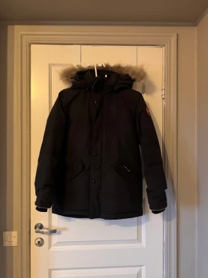 Svart dunjacka med päls från Canada Goose - Svart dunjacka från Canada Goose med avtagbar huva och fluffig pälsdetalj. Jackan har flera fickor, dragkedja och knappar framtill samt innerficka med dragkedja.