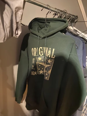 Grön hoodie med stjärntryck och huva - ENDA PÅ PLICK!!!! Mörkgrön hoodie från Original Fit med stor stjärn- och textprint framtill. Tröjan har huva med snörning och en klassisk känguruficka. Perfekt för dig som gillar en avslappnad och cool stil. Passar dig som är runt 180