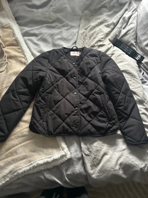 Svart quiltad jacka från Nelly XXS - Quiltad svart jacka från Nelly i storlek XXS. Jackan har rund hals, tryckknappar framtill och ett stilrent rutmönster. Perfekt för dig som gillar minimalistisk stil och vill ha en lätt jacka med snygg passform. Helt oanvänd