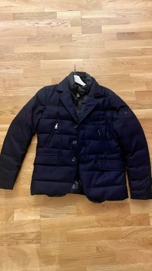 Mörkblå dunjacka från Hetregó - Snygg mörkblå dunjacka från Hetregó med kavajkrage och quiltad design. Jackan har både knappar och dragkedja framtill, flera fickor med dragkedja och en stilren metallbadge på ärmen. Insidan är fodrad med glansigt material för extra komfort och värme. Storlek 50 vilket motsvarar M