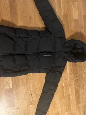 Svart lång pufferjacka Sail Racing dam - Säljer en svart lång pufferjacka från Sail Racing med huva och snygga detaljer som Sail Antarctica Expedition-patch på ärmen. Jackan har dragkedja framtill och orange zip-pullers. Perfekt för kalla dagar och har en clean, sportig look.