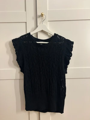 Svart stickad blus från Lindex - Säljer en svart stickad blus från Lindex i storlek S. Toppen har ett snyggt hålmönster, ribbad kant nedtill och volangdetaljer vid axlarna. Ärmlös design som ger en cool och trendig look. Perfekt att styla med jeans eller kjol!