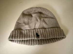 Grå mössa från Polo Ralph Lauren - Snygg grå stickad mössa från Polo Ralph Lauren med klassisk svart broderad logga framtill. Mössan har uppvikt kant med ribbad struktur och är tillverkad i mjuk ullblandning som håller dig varm. Perfekt för kalla dagar och enkel att matcha med din jacka.