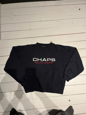 Svart sweatshirt från Chaps Ralph Lauren - Mörkblå boxig sweatshirt från Chaps Ralph Lauren med stor vit och röd logotyp på bröstet. Tröjan har rund hals, långa ärmar och ribbade muddar vid ärmslut och nederkant. Materialet är 80% bomull och 20% polyester, vilket gör den mjuk och skön.