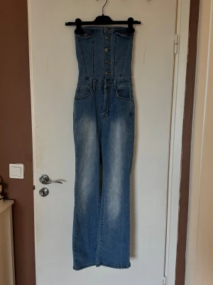 Blå jeans jumpsuit från SHEIN - Säljer en helt oanvänd blå jeans jumpsuit från SHEIN med hög midja och raka ben. Framsidan har flera knappar och markerade sömmar, medan baksidan har resår upptill och klassiska bakfickor. Materialet är jeans med lätt tvättad look. 