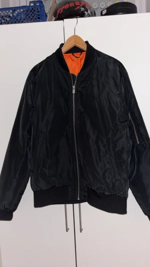 Svart bomberjacka med orange foder - Klassisk svart bomberjacka med orange foder, ribbade muddar och dragkedja framtill. Jackan har fickor på sidorna och är tillverkad i glansig polyester. Perfekt för dig som vill ha en tidlös och cool look.