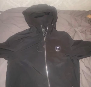 Trapstar jacka - Svart vindjacka från Trapstar med huva och dragkedja framtill. Jackan har en rund logotyp på bröstet och stor broderad Trapstar-text på ryggen. Snygg streetstil med snörning vid huvan och praktiska fickor. Perfekt för dig som gillar urban vibe.