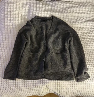 Mörkgrå kofta från Vero Moda - Mysig mörkgrå kofta från Vero Moda med två stora fickor framtill och knappar hela vägen. Koftan har långärmad passform och är tillverkad i ett stickat material som känns både chill och skönt. Perfekt att slänga på sig när det är lite kyligt ute.