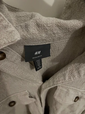 Beige overshirt från H&M Regular Fit - Snygg beige overshirt från H&M i regular fit. Skjortan har två stora bröstfickor med knappar och klassisk krage. Materialet är mjukt och har en lätt struktur, perfekt för lager-på-lager. Passar dig som gillar enkel och clean stil.
