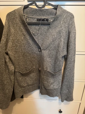 Grå stickad kofta från 157 XS - Mysig grå stickad kofta från 157 i storlek XS. Koftan har två framfickor, ribbade muddar och knappar framtill. Perfekt för lager-på-lager och chill dagar. Snygg v-ringad design som funkar till jeans eller mjukis.