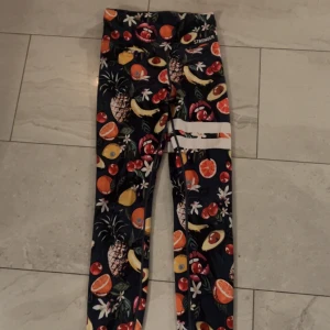 Mönstrade leggings från Stronger - Säljer ett par färgglada leggings från Stronger med fruktigt mönster i orange, gul, röd och grön på mörkblå botten. De har vita ränder på ena benet och är tillverkade i stretchigt syntetmaterial. Perfekta för träning och riktigt snygga detaljer.