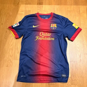 FC Barcelona Home Shirt 2012/13 – Nike Dri-FIT  - Säljer en officiell FC Barcelona matchtröja från Nike med Dri-Fit material. Tröjan är blå med röd toning i mitten, gula detaljer och klubbmärke på bröstet. Kortärmad med tryck från Qatar Foundation och UNICEF. Perfekt för dig som älskar Barca! Nästan aldrig använd och har inga skavanker! Står S men passar perfekt på M