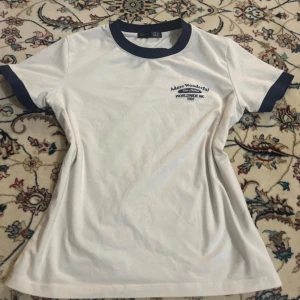 Vit t-shirt med blå detaljer DAZY - Vit t-shirt från DAZY med marinblå krage och ärmslut. Tryck på bröstet med texten 'Adore Wonderful, Worldwide Inc 1991'. Klassisk passform och tillverkad i mjuk bomull. Perfekt för en chill och stilren look.