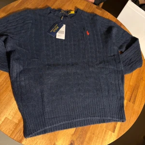 Marinblå stickad tröja Polo Ralph Lauren - Snygg marinblå stickad tröja från Polo Ralph Lauren med diskret röd logga broderad på bröstet. Tröjan har ett klassiskt kabelstickat mönster, rund hals och långa ärmar. Perfekt för dig som gillar stilrena och tidlösa plagg.