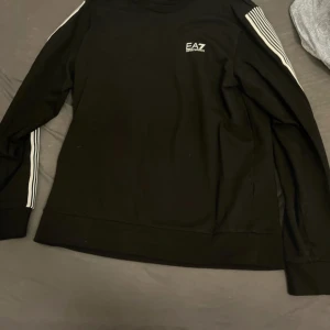 Svart långärmad tröja EA7 Armani - Svart långärmad tröja från EA7 Emporio Armani med vita ränder längs ärmarna och logga på bröstet. Tröjan har en klassisk rund hals och är gjord i mjuk bomull, perfekt för en sportig och stilren look.