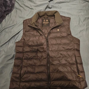 Oliv grön dunväst från Morris, medium - Snygg oliv grön dunväst från Morris i storlek medium. Västen har quiltad design, dragkedja framtill och en broderad guldfärgad lilja på bröstet. Perfekt för lager-på-lager och har en klassisk ståkrage. Materialet är dun.