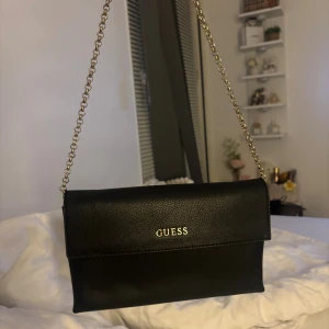 Svart axelväska från Guess - ÄKTA Snygg svart axelväska från Guess med guldfärgad kedja som axelrem och logga i guld på framsidan. Väskan har en stilren och klassisk design.💕 