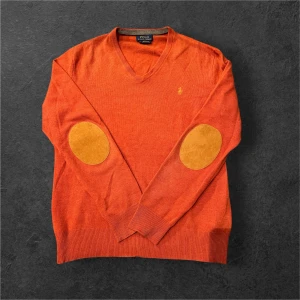 Orange v-ringad tröja Polo Ralph Lauren - Orange v-ringad tröja från Polo Ralph Lauren med klassisk gul broderad logga på bröstet. Tröjan har långa ärmar och snygga mockadetaljer på armbågarna. Perfekt för lager-på-lager och chill höststil.