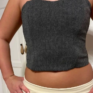 Grå tweed korsett topp ginatricot - Säljer en snygg gråmelerad korsett topp i tweed med bandeauformad överdel. Toppen är axelbandslös och har en lätt figurnära passform som ger en clean och trendig look. Perfekt att styla med jeans eller kostymbyxor för en cool vibe. Med lapp kvar på
