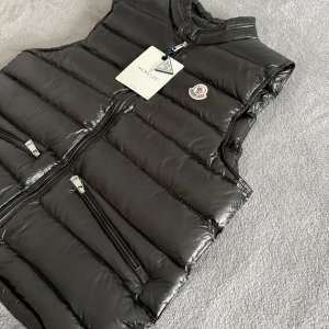 Moncler väst  - Svart, glansig dunväst från Moncler med quiltad design och ståkrage. Västen har två snedställda dragkedjefickor framtill, Moncler-logga på bröstet och en diskret logolapp på insidan. Perfekt för lager-på-lager och snygg till streetwear.