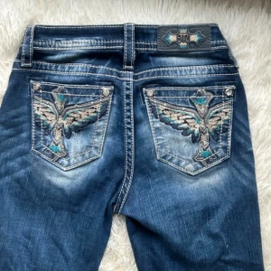 Blå bootcut Miss Me jeans  - Snygga blå jeans från Miss Me med bootcut passform. Jeansen har broderade bakfickor med turkosa och beige detaljer samt tydliga kontrastsömmar. Klassisk femficksmodell med midwaist. Perfekta för dig som vill sticka ut med coola detaljer.Använt många gånger och väldigt bra skick förutom att M:et på baksidan har rostat lite som visas på en slide. Köpt från Miss Me:s egna hemsida!!   Har varit favoritjeansen jättelänge men är tyvärr för små nu.  